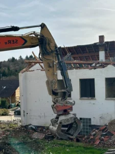 Bauphase mit der Demontage eines alten weißen Backsteingebäudes mit Ziegeldach durch einen Baggergreifer während des Rückbaus.