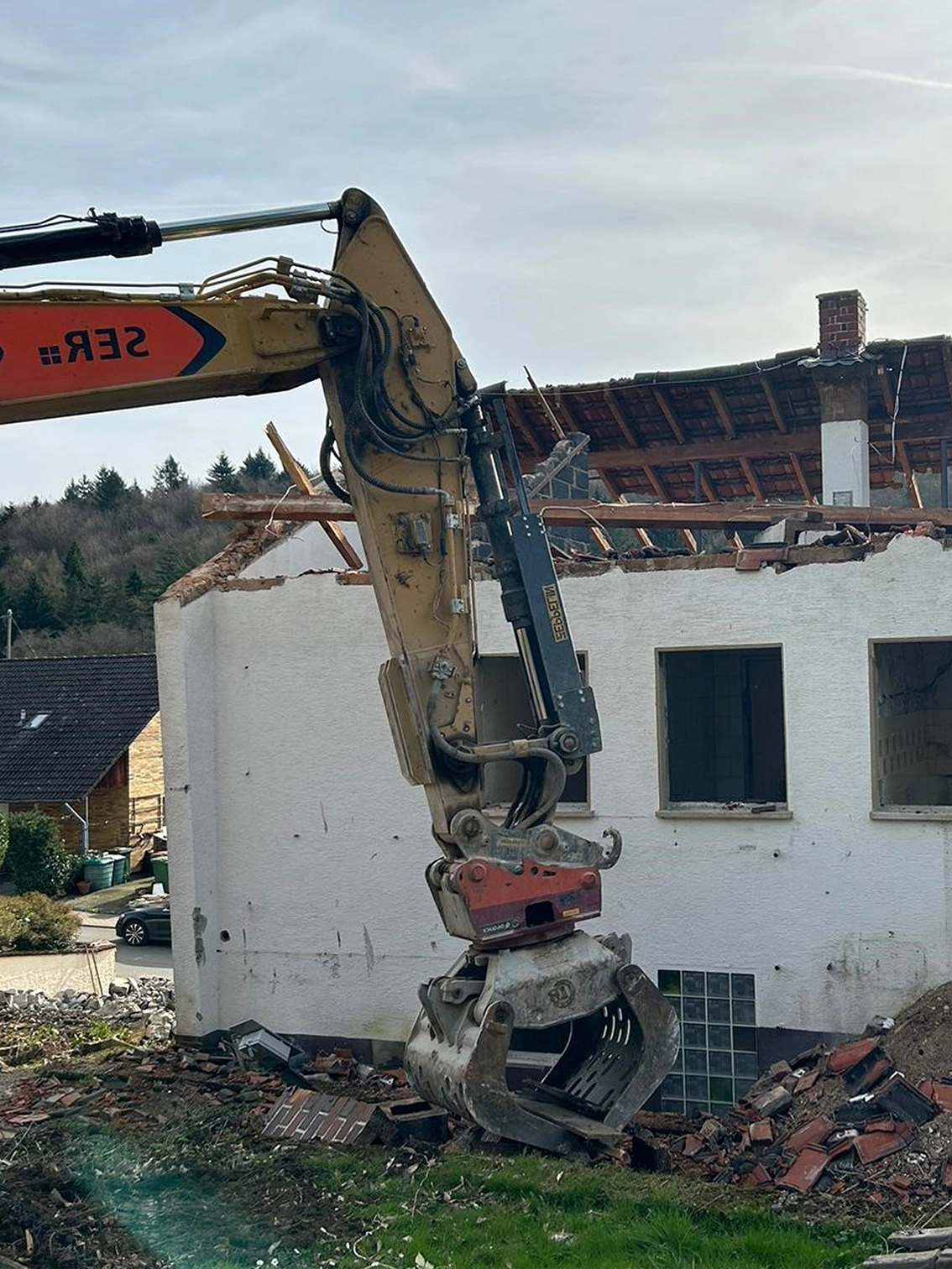 DE_United-Architektur-Demolition Bauphase mit der Demontage eines alten weißen Backsteingebäudes mit Ziegeldach durch einen Baggergreifer während des Rückbaus.