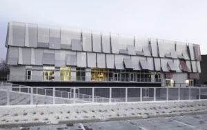 Weite Außenansicht eines dreistöckigen institutionellen Gebäudes, das in eine dynamische, mehrlagige Hülle aus silbernen Mesh-Paneelen und Glasfenstern über einem umzäunten Gelände gehüllt ist.