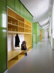 Heller Kindergartenflur mit langen, integrierten Garderoben aus gelbem Holz vor leuchtend grünen, transluzenten Wandpaneelen.