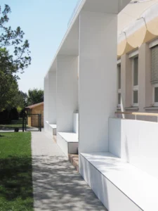 Architekturfotografie der Außenansicht einer Bildungseinrichtung, die einen langen weißen modularen Pavillon mit integrierten Sitzbänken entlang eines gepflasterten Weges zeigt.