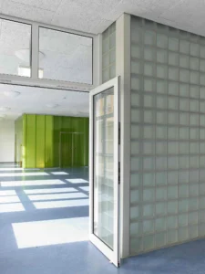 Innenansicht einer Bildungseinrichtung mit einem weißen Türrahmen neben einer Wand aus mattierten Glasbausteinen, im Hintergrund sind grüne Wandpaneele erkennbar.