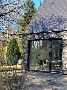 Sonnige Außenansicht einer modernen Hausecke mit dunkler Holzverkleidung, steilem Giebeldach und einem großen Schiebefenster, das einen Blick in den Essbereich freigibt.