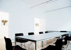 Innenansicht eines geräumigen Kanzlei-Konferenzraums mit einem langen, minimalistischen weißen Besprechungstisch, schwarzen Ledersesseln und elegantem Fischgrätparkett.
