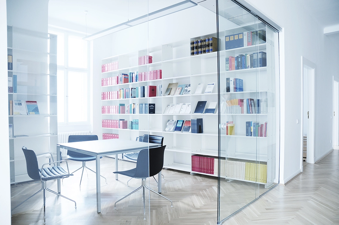 DE_United-Architektur-Kanzlei-Meyer-Köring-Berlin-Umbau-Möbel-office1 Modernes Büro-Innendesign mit einem Konferenzraum, der durch eine rahmenlose Glastrennwand abgetrennt ist, ausgestattet mit minimalistischen weißen Regalen und klassischem Holzparkett.