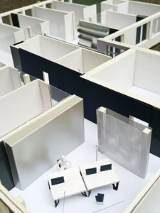 Architekturmodell des Büro-Grundrisslayouts, das weiße Miniaturschreibtische, eine Trennwand aus Mesh, dunkle Wandpaneele und die räumliche Aufteilung das einzelnen Räume zeigt.