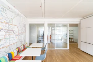 Pausenraum im Büro mit kleinen weißen Tischen, blauen Kunststoffstühlen, einer bunt gestreiften Sitzbank und einer Wand, die vollständig mit einer großen Grafik des Berliner Nahverkehrsnetzes bedeckt é, neben Glastrennwänden.