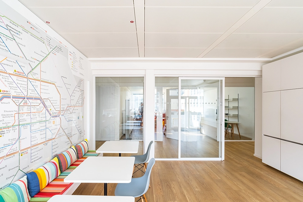 DE_United-Architektur-Ogletree-Deakins-Law-Firm-Office2 Pausenraum im Büro mit kleinen weißen Tischen, blauen Kunststoffstühlen, einer bunt gestreiften Sitzbank und einer Wand, die vollständig mit einer großen Grafik des Berliner Nahverkehrsnetzes bedeckt é, neben Glastrennwänden.