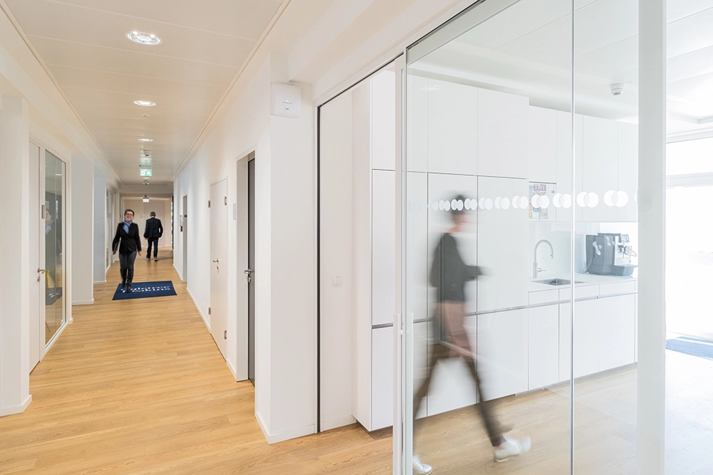 DE_United-Architektur-Ogletree-Deakins-Law-Firm-Office3 Langer Büroflur mit hellem Eichenparkett und weißen Wänden, mit Blick vorbei an einer transparenten Glastrennmand, die eine minimalistische weiße Teeküche offenbart, während Personen in Bewegungsunschärfe vorbeigehen