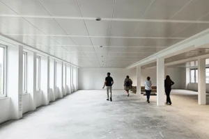 Eine Gruppe von vier Personen läuft durch einen großen, völlig leeren Rohbaubüroraum mit unbehandeltem Betonboden, einer Rasterdecke und einer langen Fensterfront vor der Sanierung.