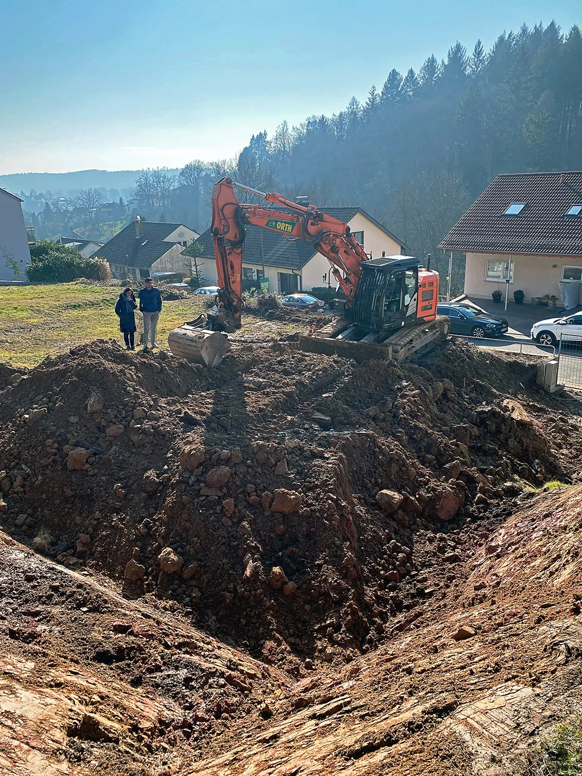 DE_United-Architektur-Start-Construction-House Grundstücksvorbereitung und Erdarbeiten auf einem steilen Wohngrundstück, mit einem orangenen Kettenbagger beim Erdaushub für o Fundament am Hang.