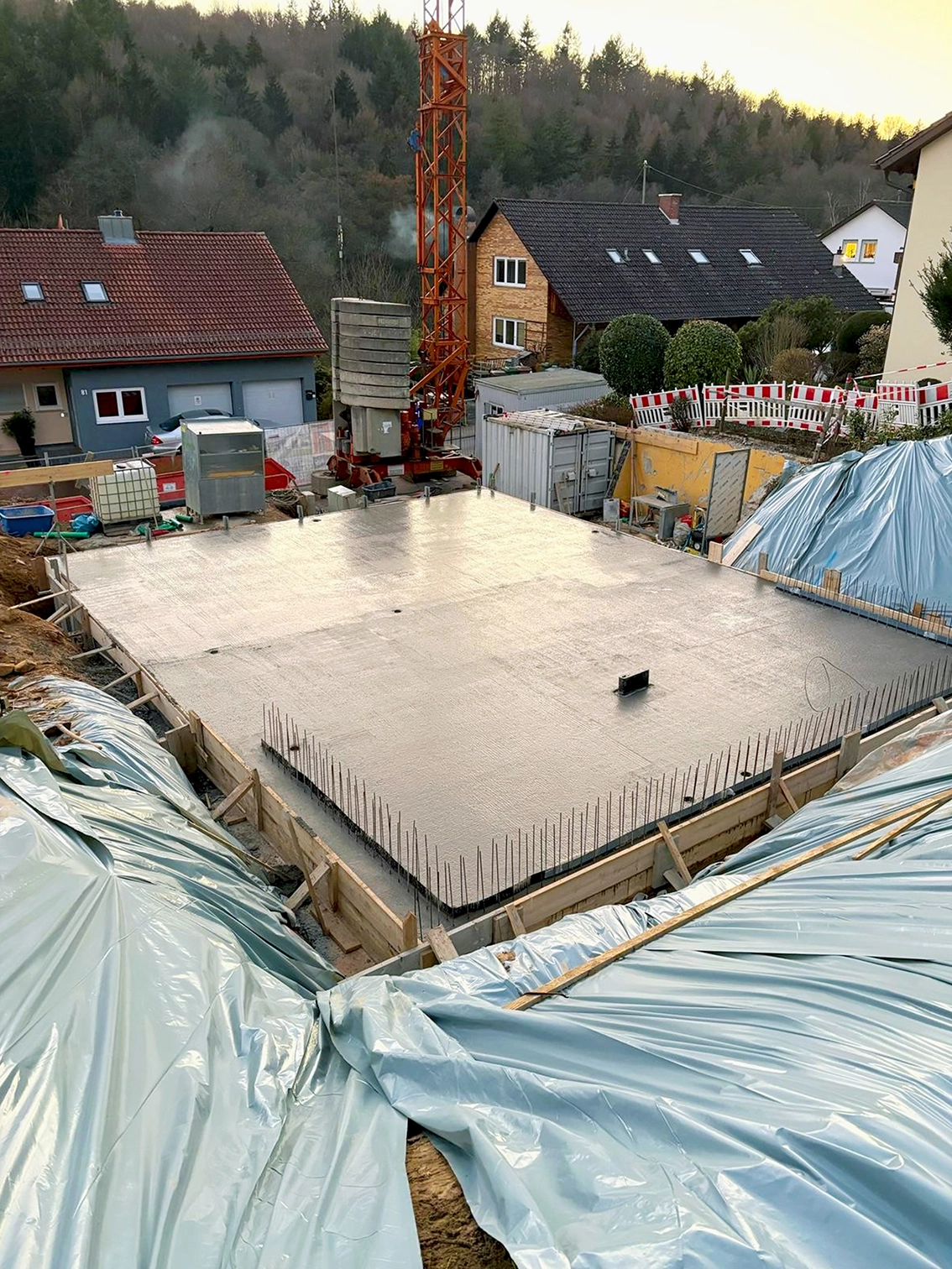 DE_United-Architektur-Start-Construction-House3 Gefilmte Draufsicht auf eine frisch gegossene Betonbodenplatte mit vertikalen Anschlussbewehrungen auf einer Hangbaustelle.