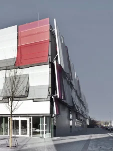 Architekturaufnahme mit Blick nach oben auf eine moderne Gebäudefassade, die eine Reihe von angewinkelten, transluzenten Mesh-Sonnenschutzelementen sowie ein einzelnes, markantes rotes Paneel nahe der Ecke zeigt.