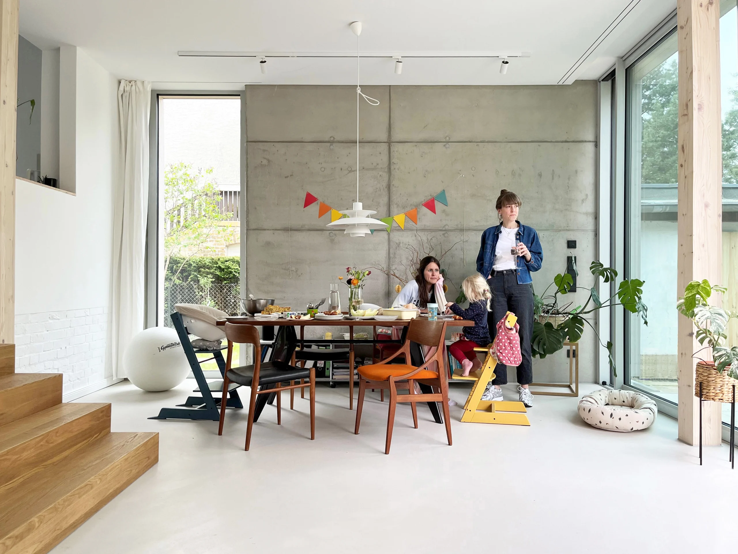 DE_United-Architektur-Yew Tree Living-dining-area Moderne Esszimmer-Inneneinrichtung mit einer Sichtbetonwand, minimalistischen Designmöbeln, großen bodentiefen Fenstern und hellem, offenem Raumkonzept.