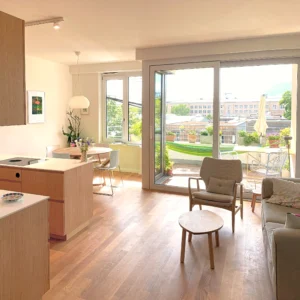 Bright open-concept apartment interior showing a modern wooden kitchen island, a living area with an armchair and sofa, and large sliding glass doors opening to a sunlit balcony terrace.