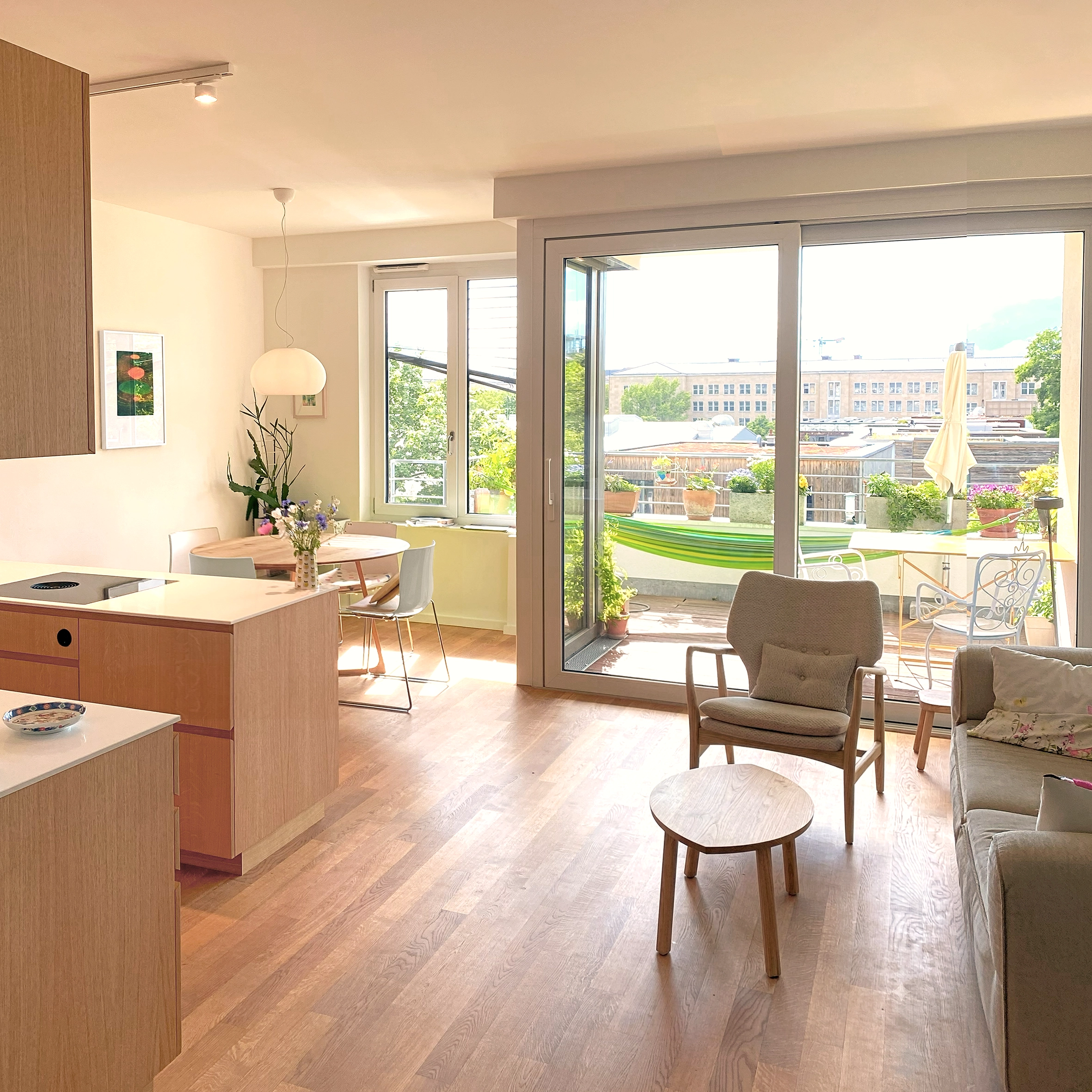 Bright open-concept apartment interior showing a modern wooden kitchen island, a living area with an armchair and sofa, and large sliding glass doors opening to a sunlit balcony terrace.