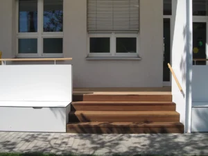 Architectural detail shot of an outdoor wooden terrace entry staircase with a minimalist white side bench, wooden handrails, and large windows with integrated blinds.
