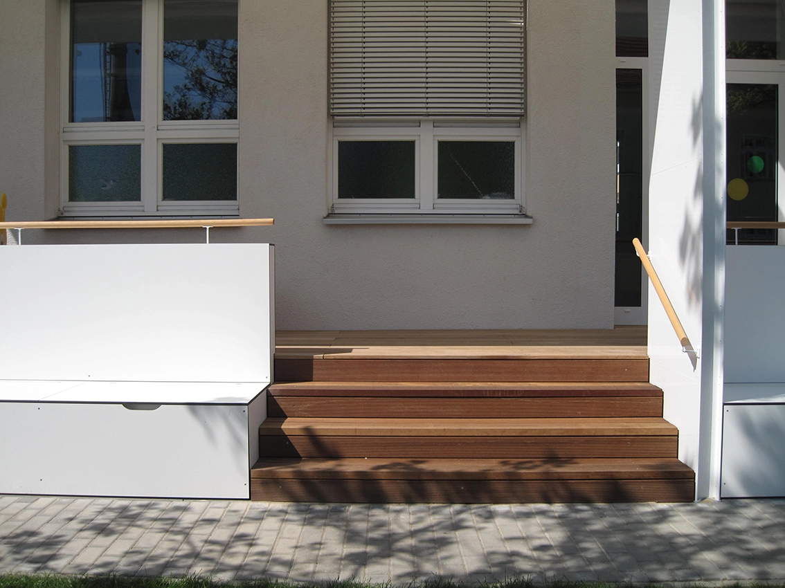 EN_United-Architektur-Haus-des-Kindes-Nursery-school-Außentreppe Architectural detail shot of an outdoor wooden terrace entry staircase with a minimalist white side bench, wooden handrails, and large windows with integrated blinds.