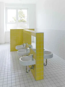 Child-friendly bathroom design showcasing low-mounted small washbasins installed on a central divider wall tiled in bright yellow.