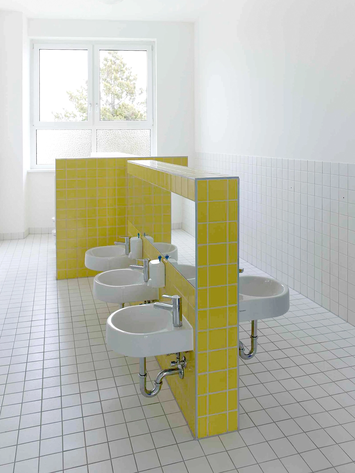EN_United-Architektur-Haus-des-Kindes-Nursery-school-Badezimmer Child-friendly bathroom design showcasing low-mounted small washbasins installed on a central divider wall tiled in bright yellow.