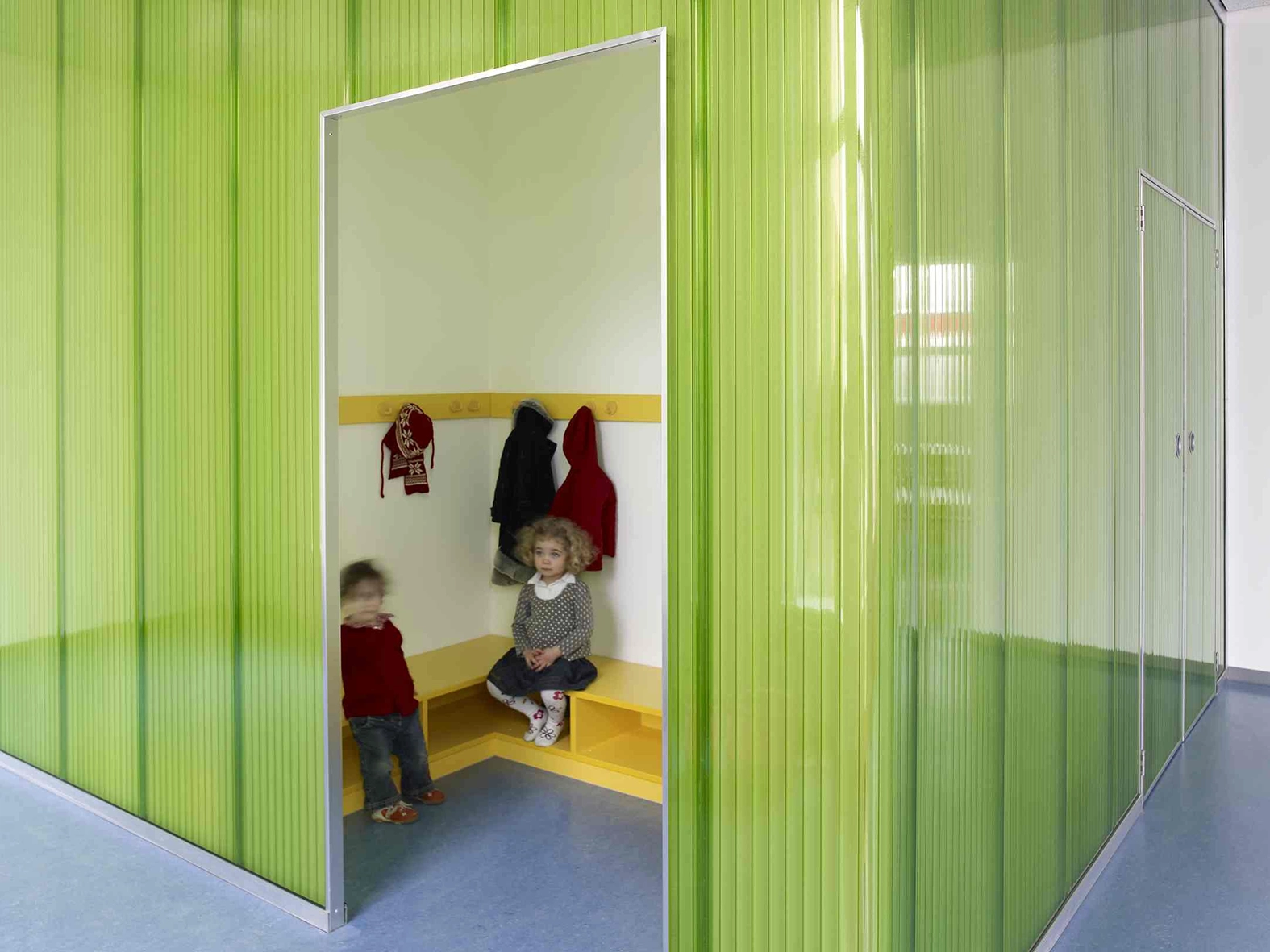 EN_United-Architektur-Haus-des-Kindes-Nursery-school-Garderobe Small cloakroom niche for children with integrated yellow benches and coat hooks, framed by a corrugated green translucent partition.