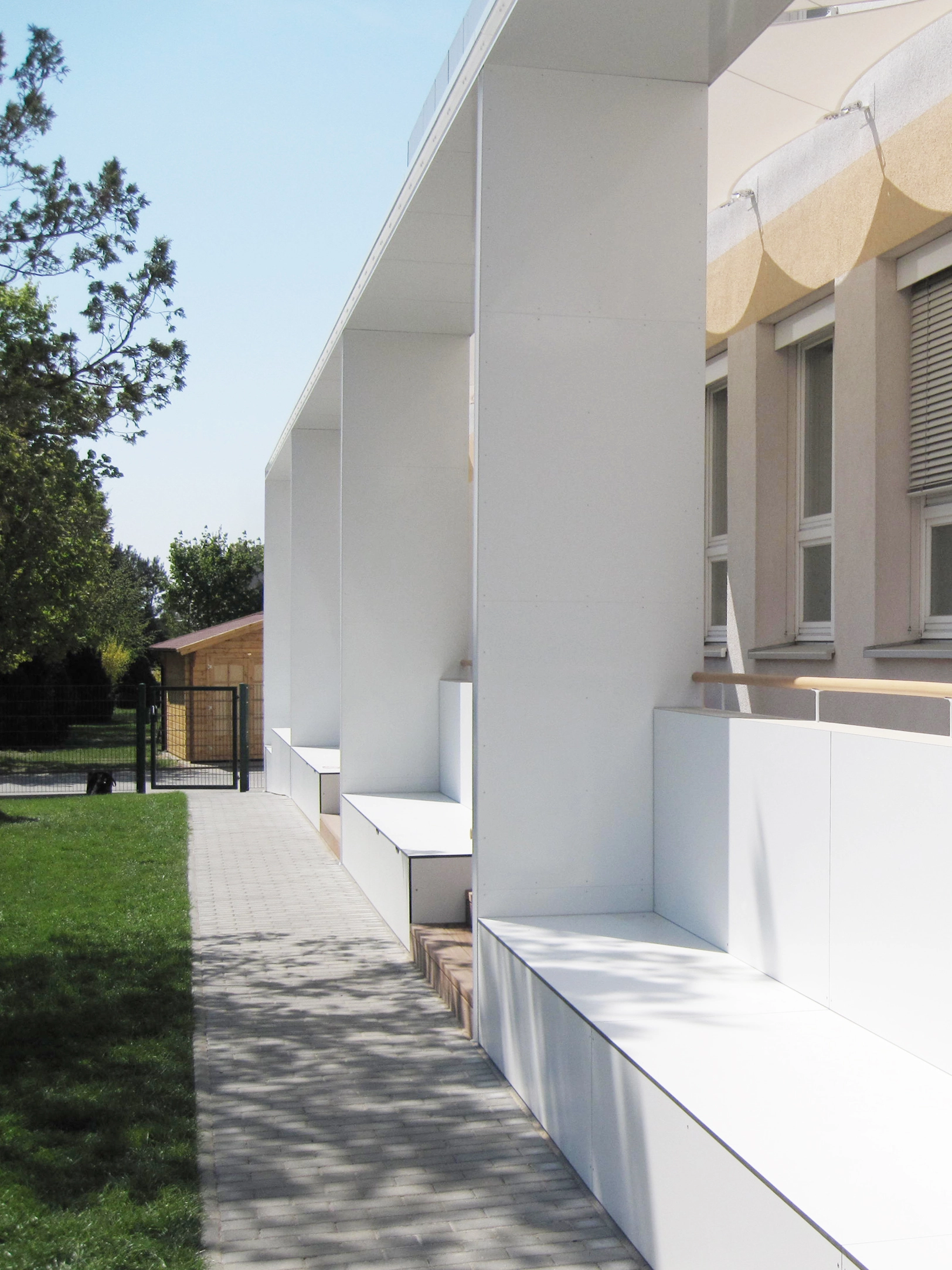 Architectural photograph of an educational facility exterior showing a long white modular structural pavilion with integrated outdoor seating benches along a stone path.