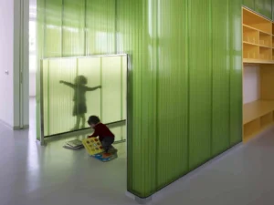 Kindergarten interior design featuring bright green translucent corrugated partitions, custom yellow shelving units, and a child playing with a book.