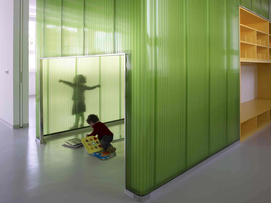 EN_United-Architektur-Haus-des-Kindes-Nursery-school-Nische Kindergarten interior design featuring bright green translucent corrugated partitions, custom yellow shelving units, and a child playing with a book.