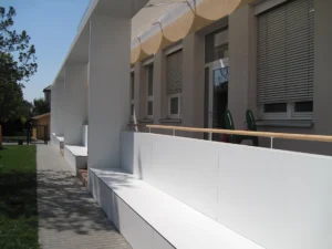 Architectural photograph of an educational facility exterior showing a long white modular structural pavilion with integrated outdoor seating benches along a stone path.
