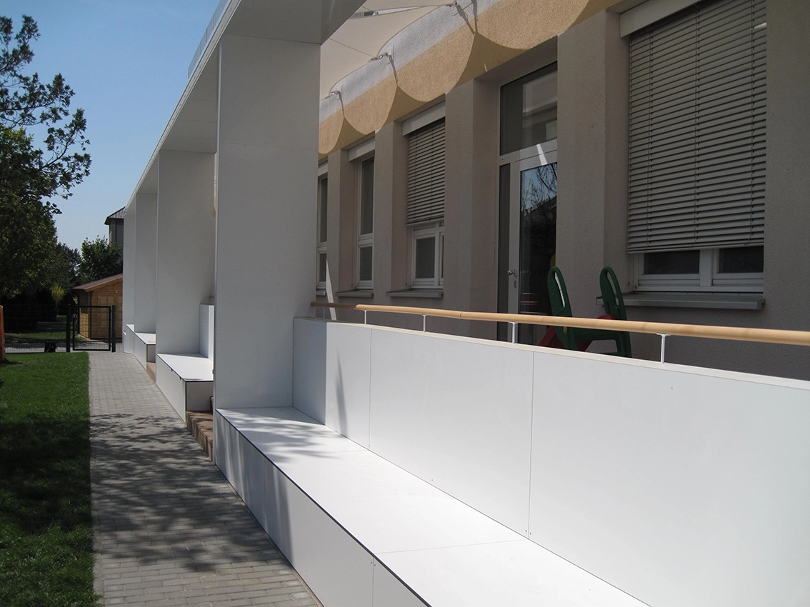 EN_United-Architektur-Haus-des-Kindes-Nursery-school-Sitzbank1 Architectural photograph of an educational facility exterior showing a long white modular structural pavilion with integrated outdoor seating benches along a stone path.