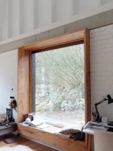 Large square window with a deep natural oak frame used as a window seat, set in a white brick wall next to a minimalist home office desk.