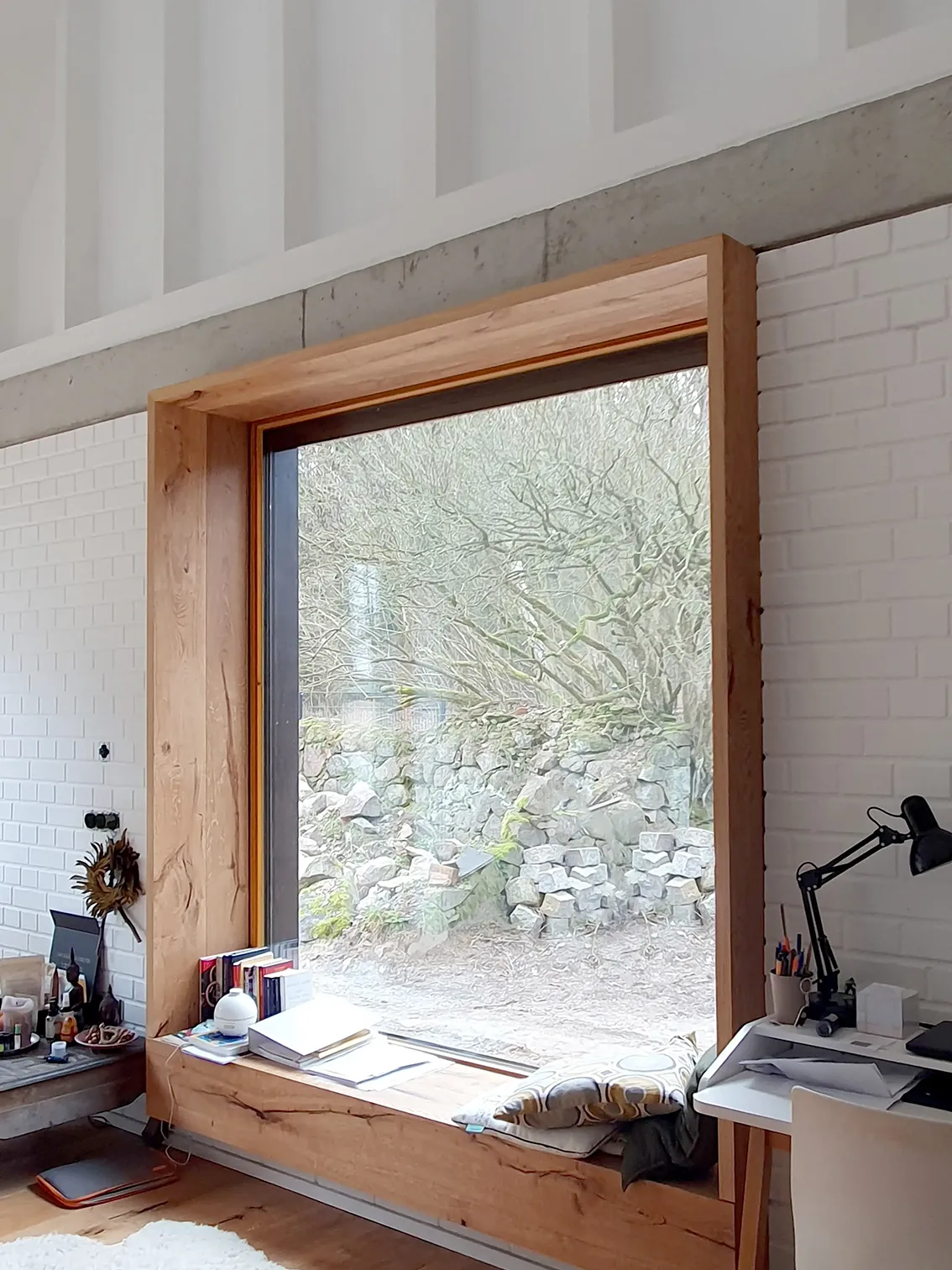 Large square window with a deep natural oak frame used as a window seat, set in a white brick wall next to a minimalist home office desk.