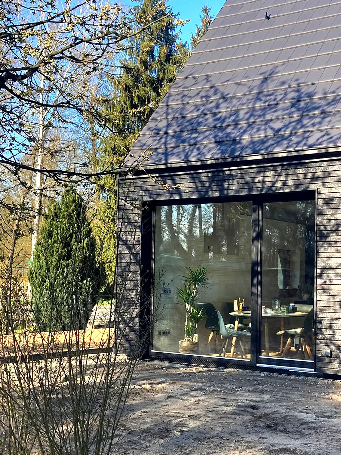 EN_United-Architektur-House-Uckermark-Lakes-Nature-Park-Garden Sunny exterior view of a modern house corner featuring dark timber cladding, a steep gabled roof, and a large sliding window showcasing a glimpse of the dining interior.