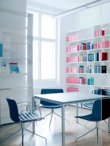Bright law firm meeting room interior with a white minimalist table, dark blue office chairs, herringbone parquet flooring, and a large white bookshelf filled with legal binders.