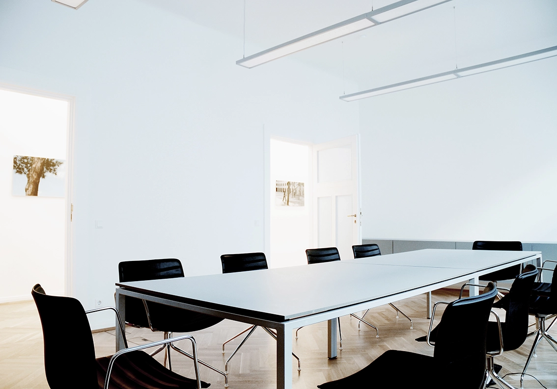 EN_United-Architektur-Kanzlei-Meyer-Köring-Berlin-Umbau-Möbel-meeting-room Interior view of a spacious law firm conference room featuring a long minimalist white meeting table, black leather chairs, and elegant herringbone parquet flooring.