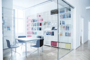Modern office interior design showing a conference room enclosed by a seamless glass partition wall, featuring minimalist white shelving and classic wooden parquet flooring.