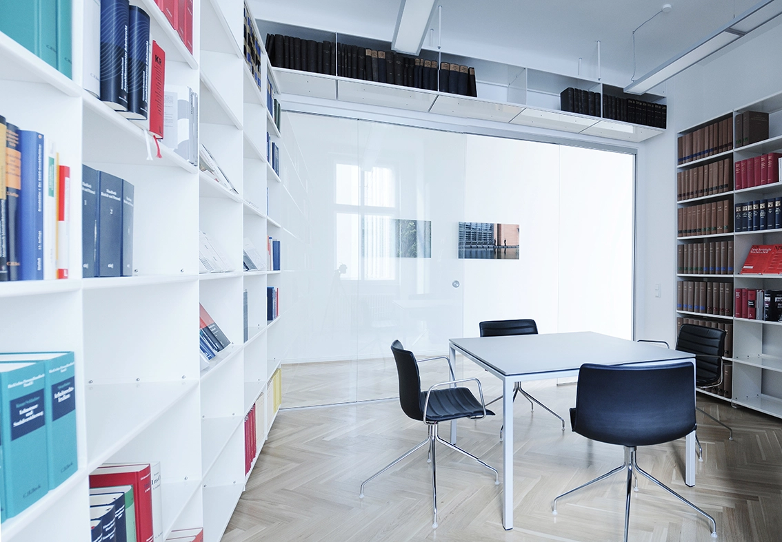 EN_United-Architektur-Kanzlei-Meyer-Köring-Berlin-Umbau-Möbel-office2 Spacious corporate meeting room with white custom library shelving spanning across the walls, black leather swivel chairs, and sleek glass partitions under a bright white ceiling.