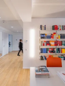 Bright corporate office interior showing a long white meeting table with vibrant orange chairs in front of a white wall shelf filled with colorful law books, opening into a wide wooden floor corridor.
