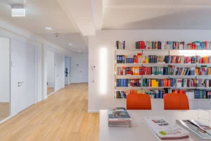 Bright corporate office interior showing a long white meeting table with vibrant orange chairs in front of a white wall shelf filled with colorful law books, opening into a wide wooden floor corridor.