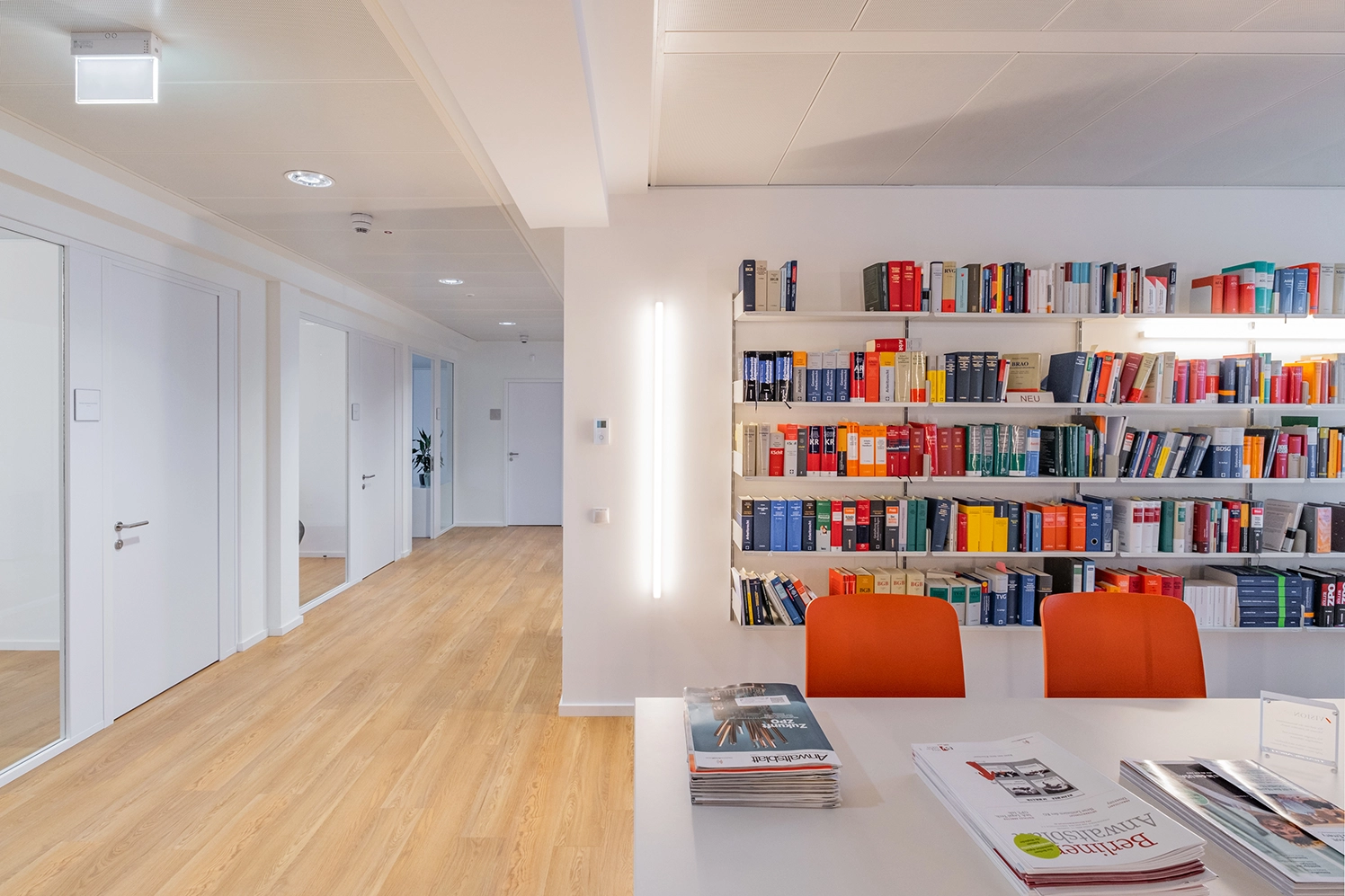 Bright corporate office interior showing a long white meeting table with vibrant orange chairs in front of a white wall shelf filled with colorful law books, opening into a wide wooden floor corridor.