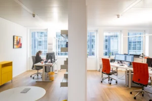 Bright, modern office interior divided by a central white partition wall, showing a woman working at a desk on the left and empty workstations with vibrant orange office chairs on the right.