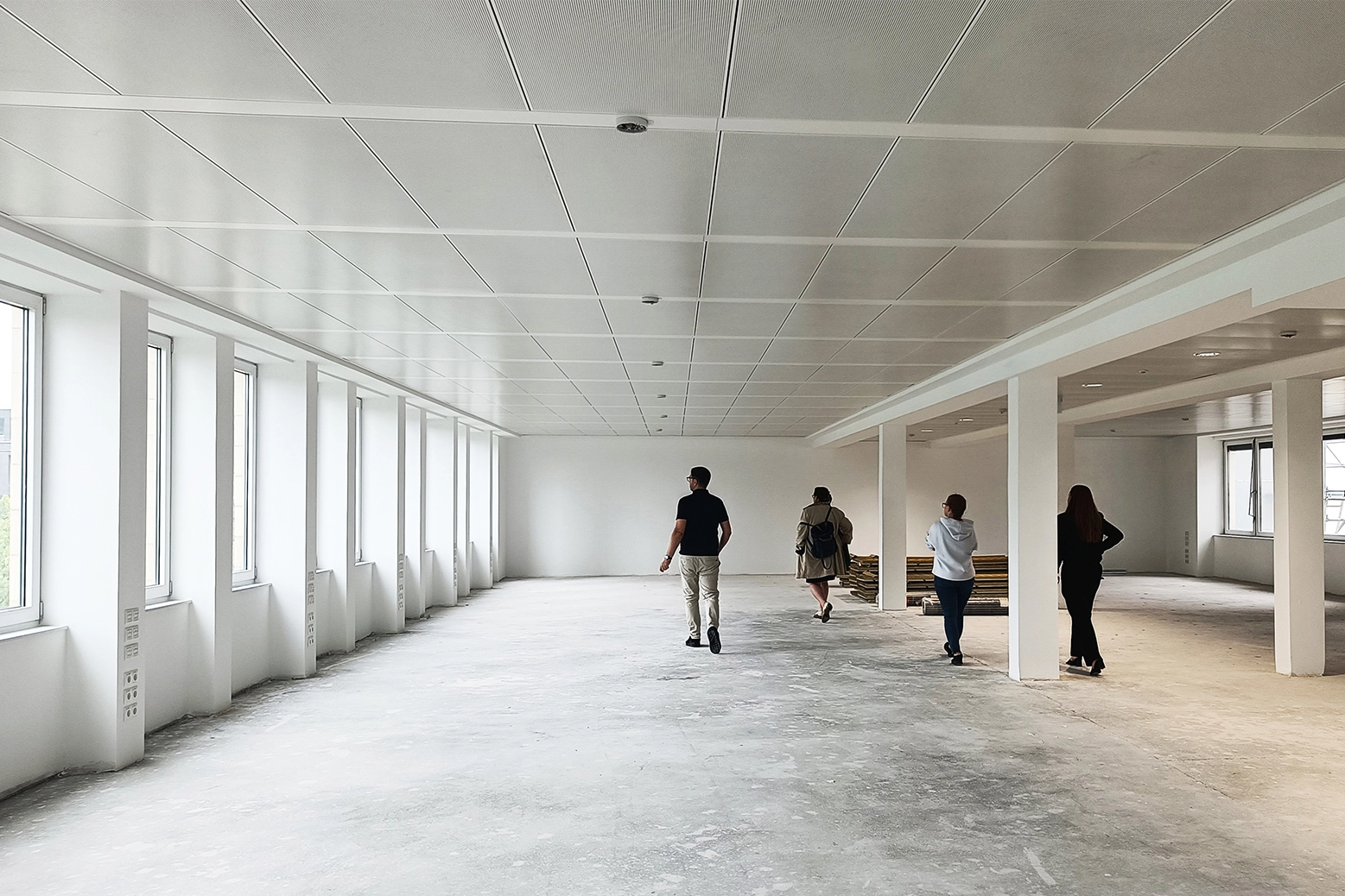 EN_United-Architektur-Ogletree-Deakins-Law-Firm-Office5 Group of four people walking through a large, completely empty raw office space with unfinished concrete floors, a grid ceiling, and a long row of windows prior to renovation.