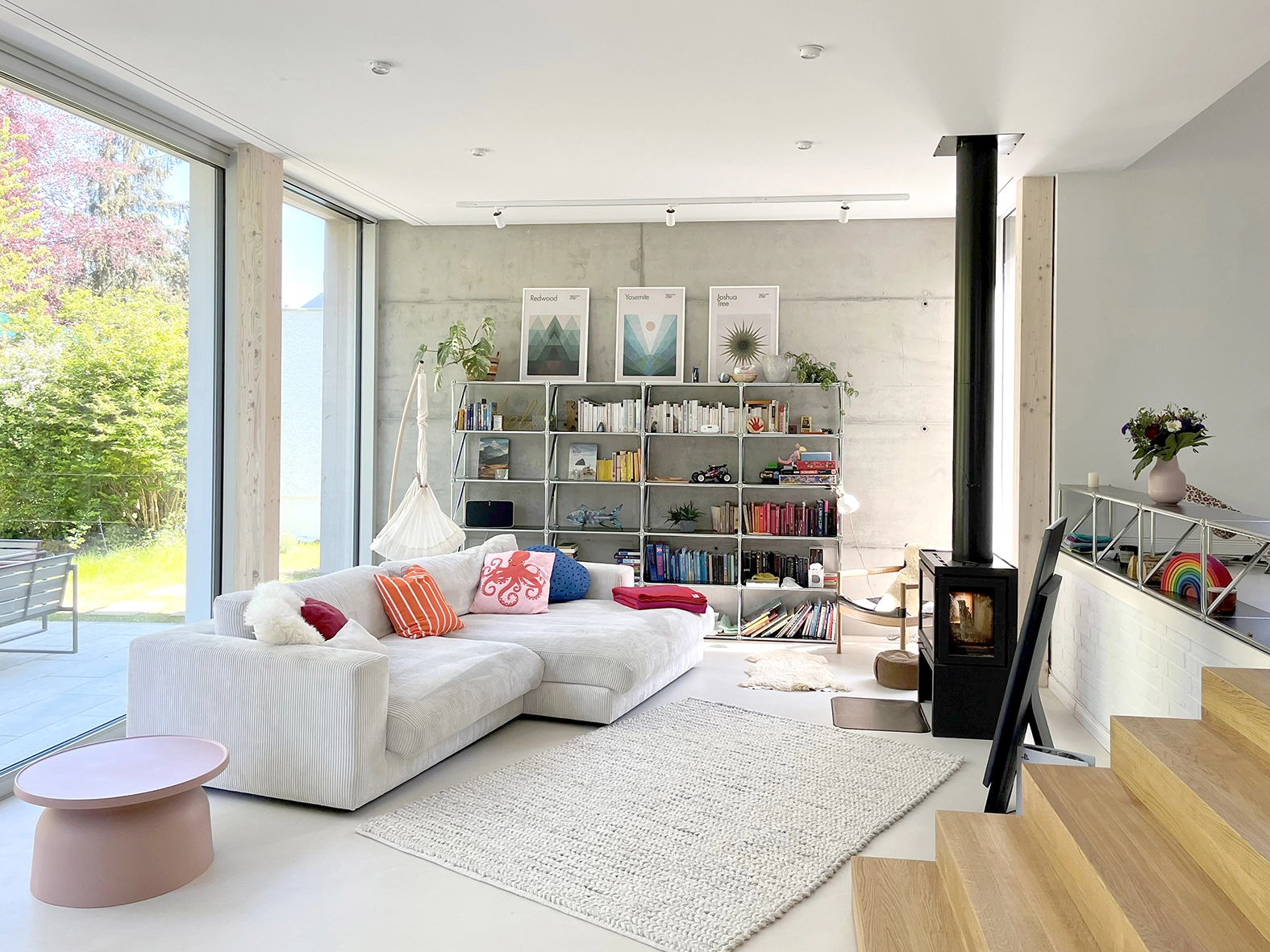 Modern open-plan living room with an exposed concrete wall, freestanding black fireplace stove, modular white sofa, and large floor-to-ceiling glass windows.