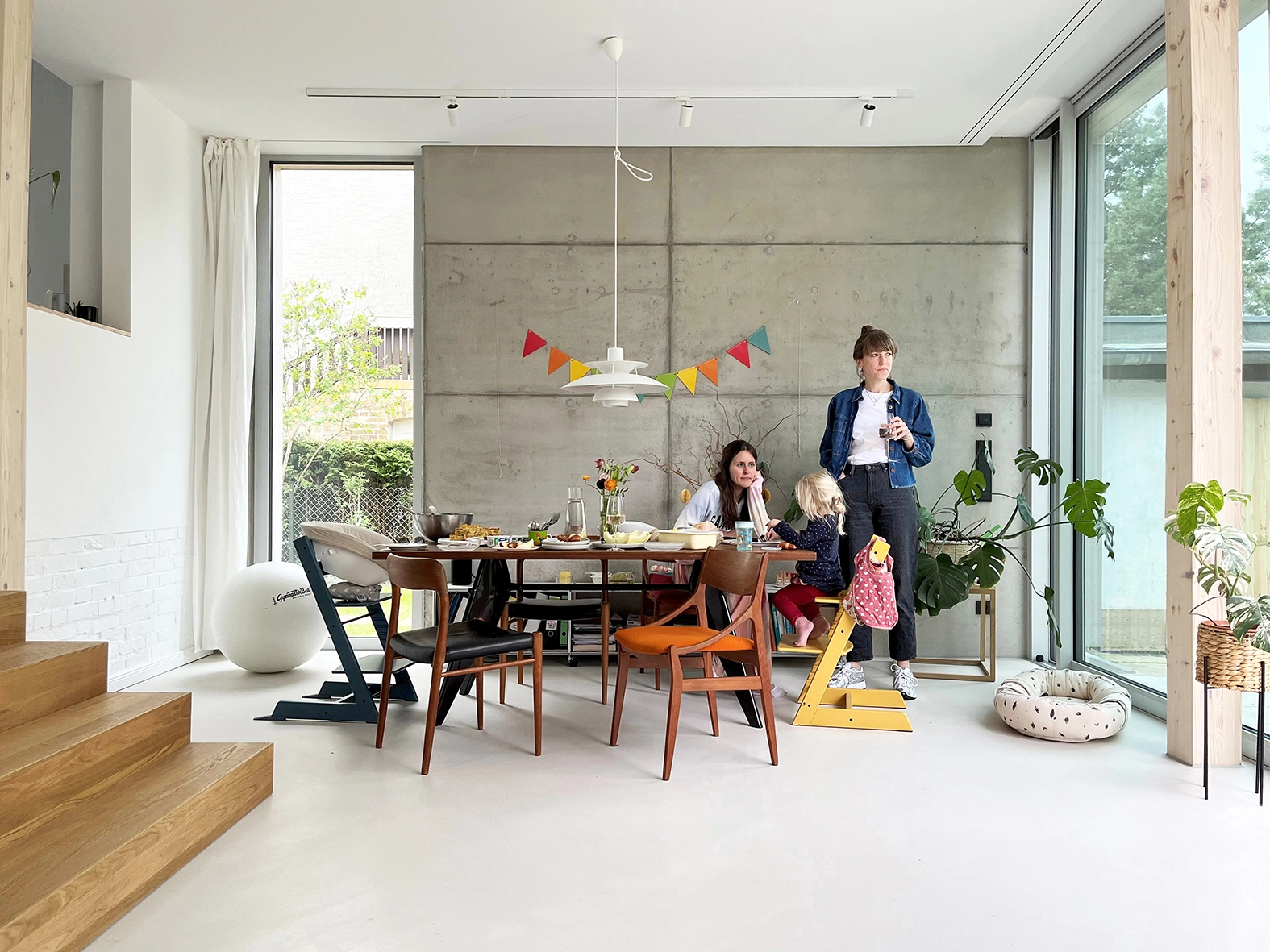 EN_United-Architektur-Yew Tree Living-dining-area Modern dining room interior featuring an exposed concrete wall, minimalist design furniture, large floor-to-ceiling windows, and bright open space.