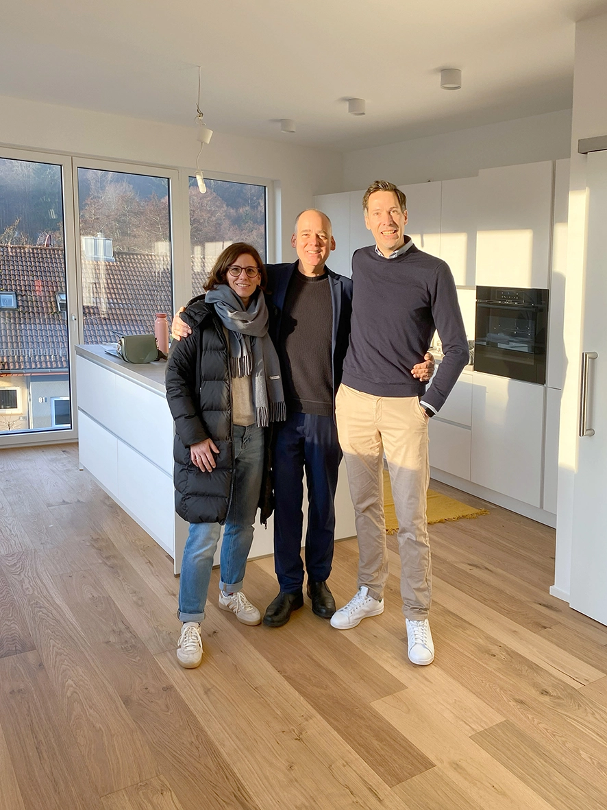 Three people smiling inside a modern kitchen and living space featuring lightweight oak flooring, minimalist white cabinets, and large floor-to-ceiling windows with a hillside view.