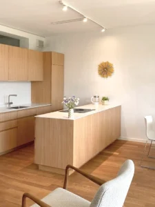 Modern minimalist kitchen design featuring light oak wood cabinetry, a long central island with a built-in cooktop, hardwood flooring, and a decorative gold sunburst wall clock.