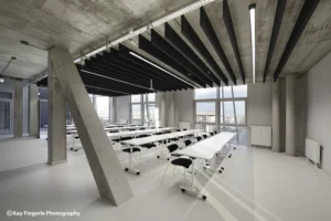 Bright seminar room with rows of modular white training tables, black chairs, an exposed concrete structural column, and acoustic ceiling panels.