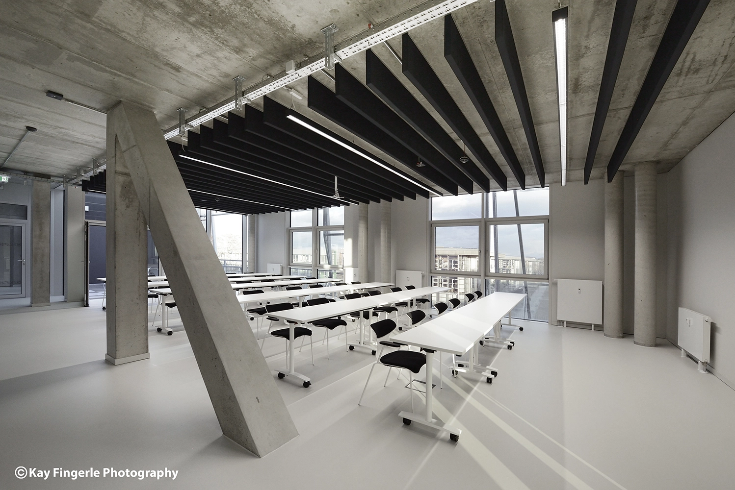 Bright seminar room with rows of modular white training tables, black chairs, an exposed concrete structural column, and acoustic ceiling panels.