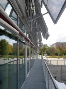 Detailed architectural view along an outdoor steel grated balcony serving the FabLab, highlighting the open glass doors and the mechanical armature of the translucent facade shading panels.