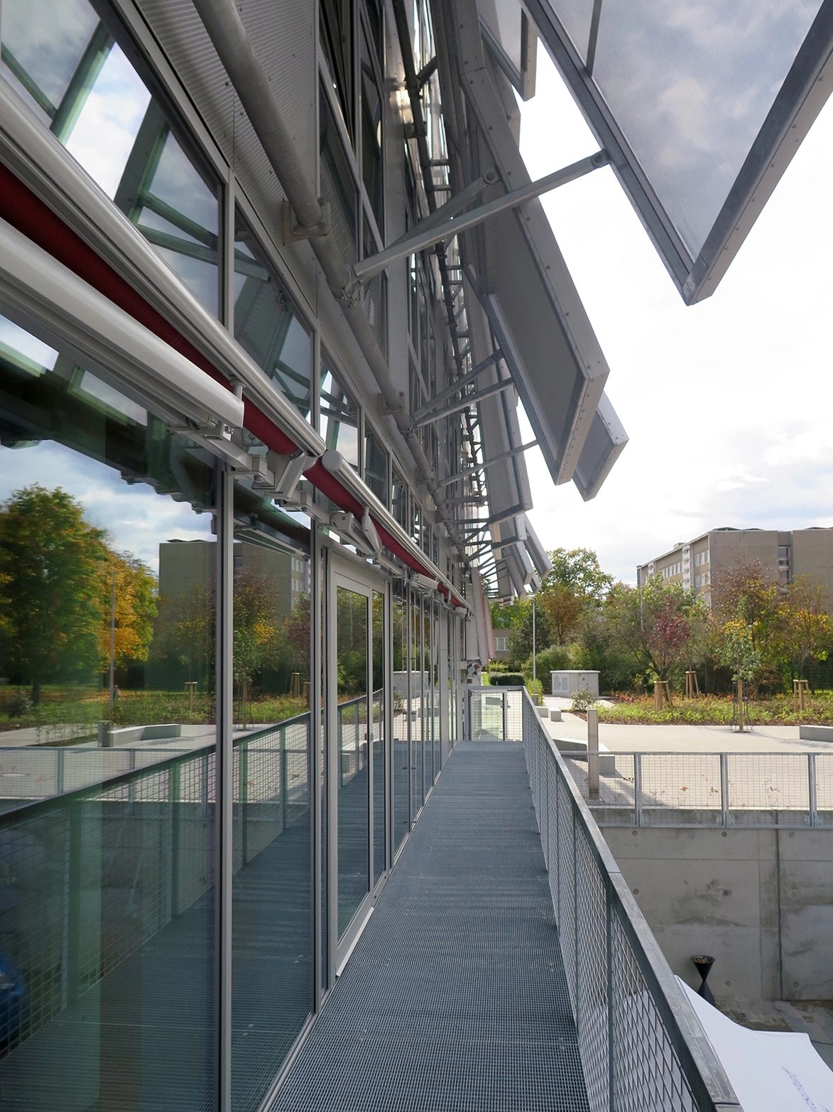 Detailed architectural view along an outdoor steel grated balcony serving the FabLab, highlighting the open glass doors and the mechanical armature of the translucent facade shading panels.